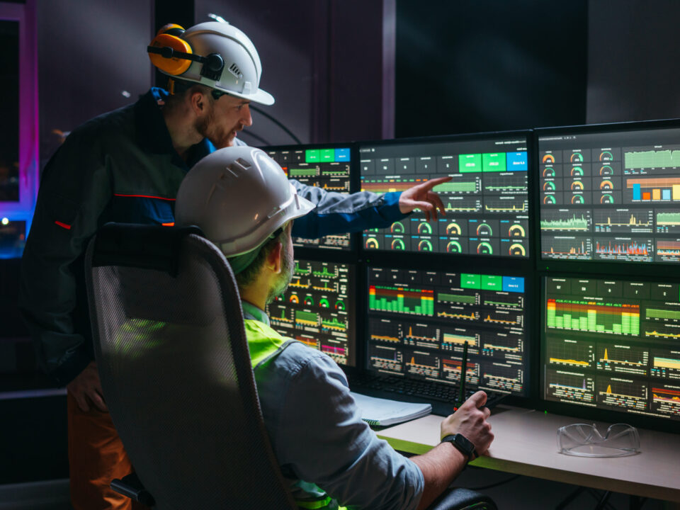 Head engineer and factory operator wearing safety vests and hard hats wollowing product process on factory uses SCADA system and industry 4.0. Two operators controll assembly line using screens with human-machine interface